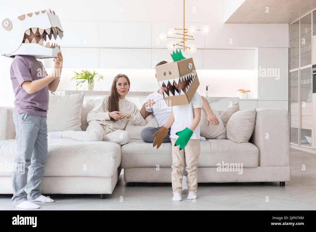 Glückliche Familie von Eltern und zwei Kindern, die zu Hause Dinosaurier spielen, Kinder tragen handgefertigte Kostümmaske aus Pappe Stockfoto