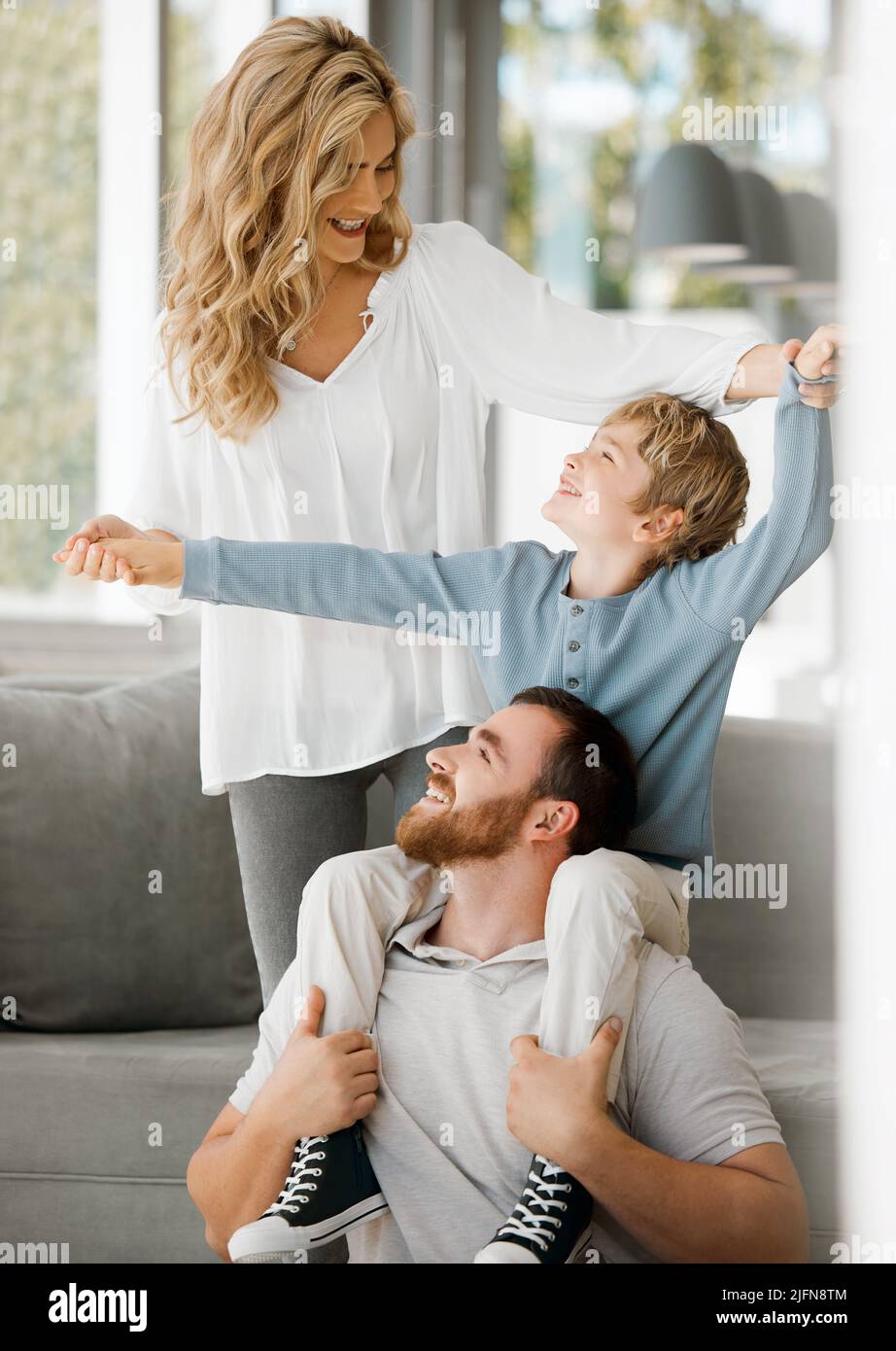 Glückliche kaukasische Familie, die sich zu Hause anfreute. Aufgeregter kleiner Junge, der auf den Schultern seines Vaters sitzt und die Hände seiner Mutter hält, während er vorgibt, für ihn zu fliegen Stockfoto