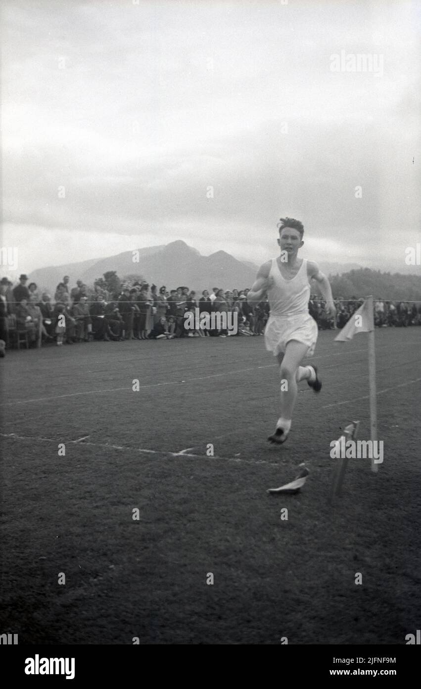 1952, historisch, ein älterer männlicher Student in Weste und Shorts, der auf einer Grasbahn auf einem Sportplatz läuft, der kurz vor der Ziellinie steht, beobachtet von Eltern und anderen Schulkindern auf der Seite, Highlands, Schottland, Großbritannien. Stockfoto