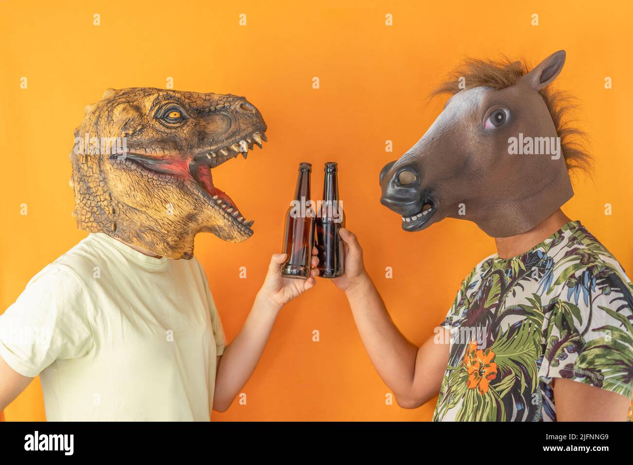 Lustige paar Freunde, die Spaß haben, Bierflaschen zu toasten, tragen eine Pferd- und Dinosaurier-Tiermaske auf orangefarbenem Hintergrund isoliert. Stockfoto
