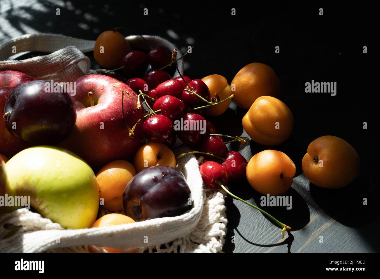 Frische reife Früchte und Beeren auf schwarzem Holzhintergrund in einem ökologischen Netzbeutel. Lifestyle. Harte Schatten.Sonnenlicht. Speicherplatz kopieren. Stockfoto