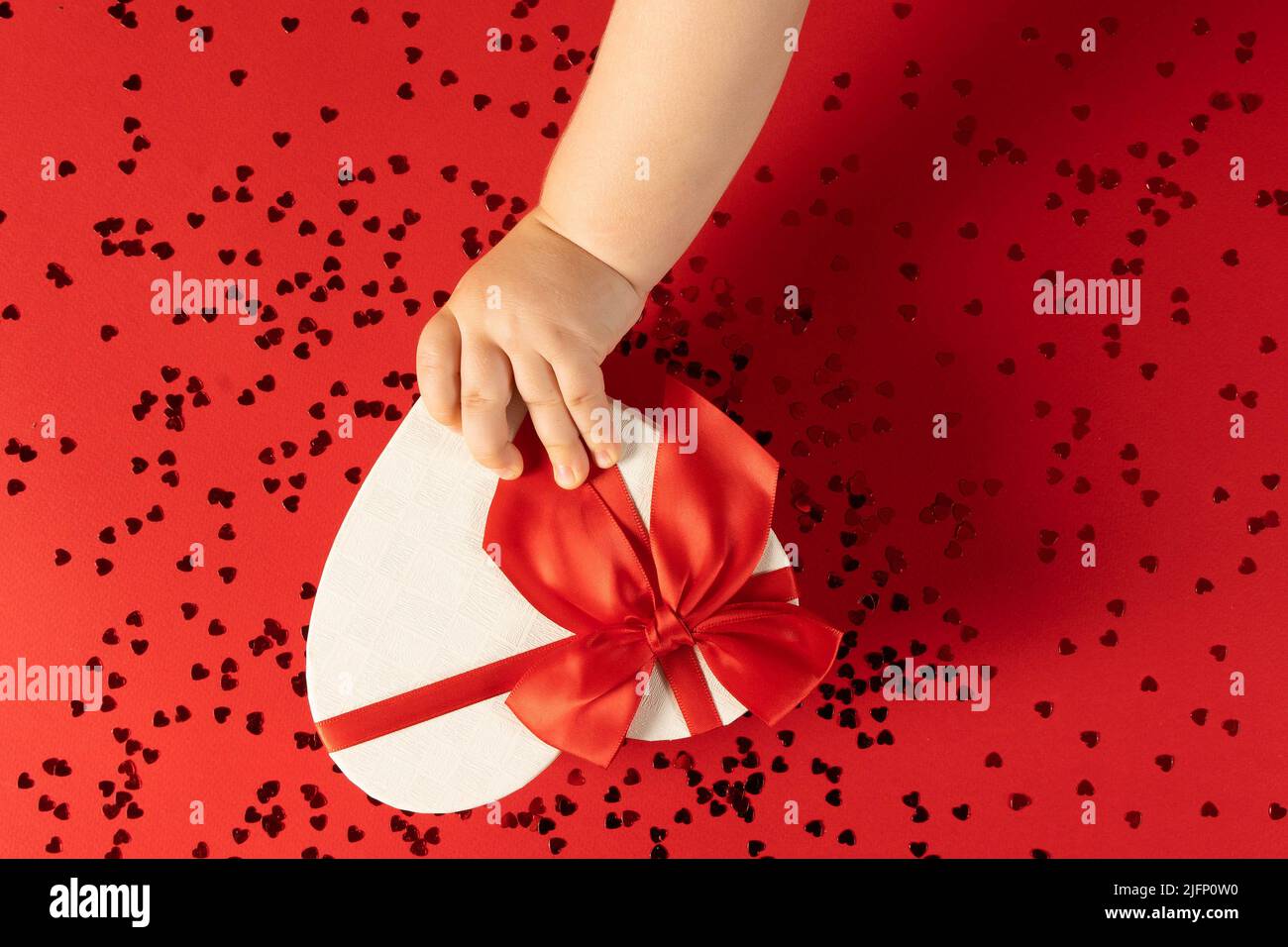 Foto von Valentinstag Dekoration von oben weiße herzförmige Geschenkbox mit roten Satinband Schleife und Konfetti auf rotem Hintergrund.Eine Grußkarte. Stockfoto