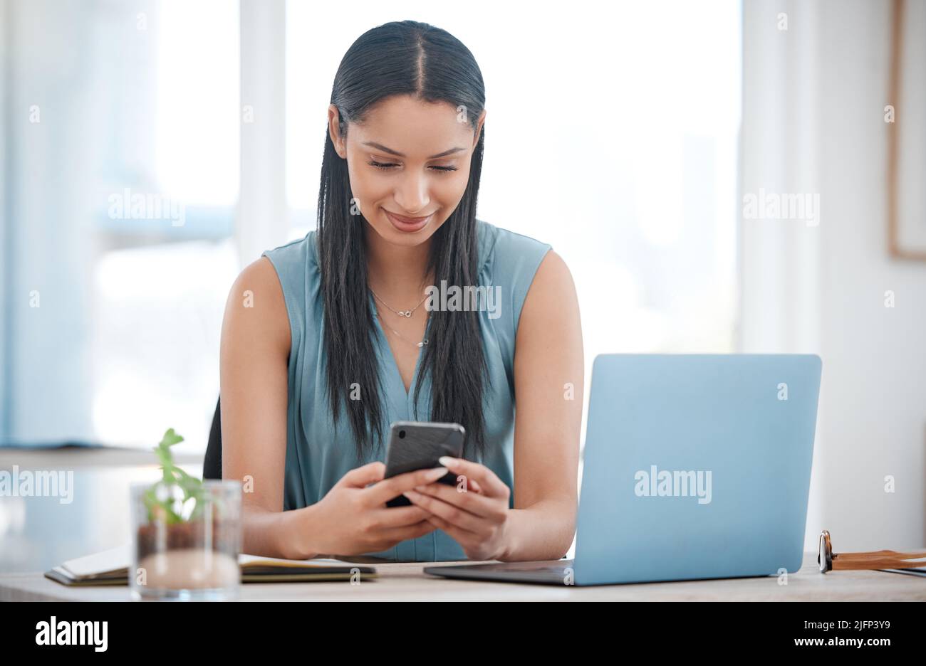 Glückliche junge Geschäftsfrau mit gemischtem Rennen lächelt, während sie ein Telefon benutzt und an einem Laptop arbeitet, der in einem Bürostuhl sitzt. Eine hispanische Chefin Stockfoto