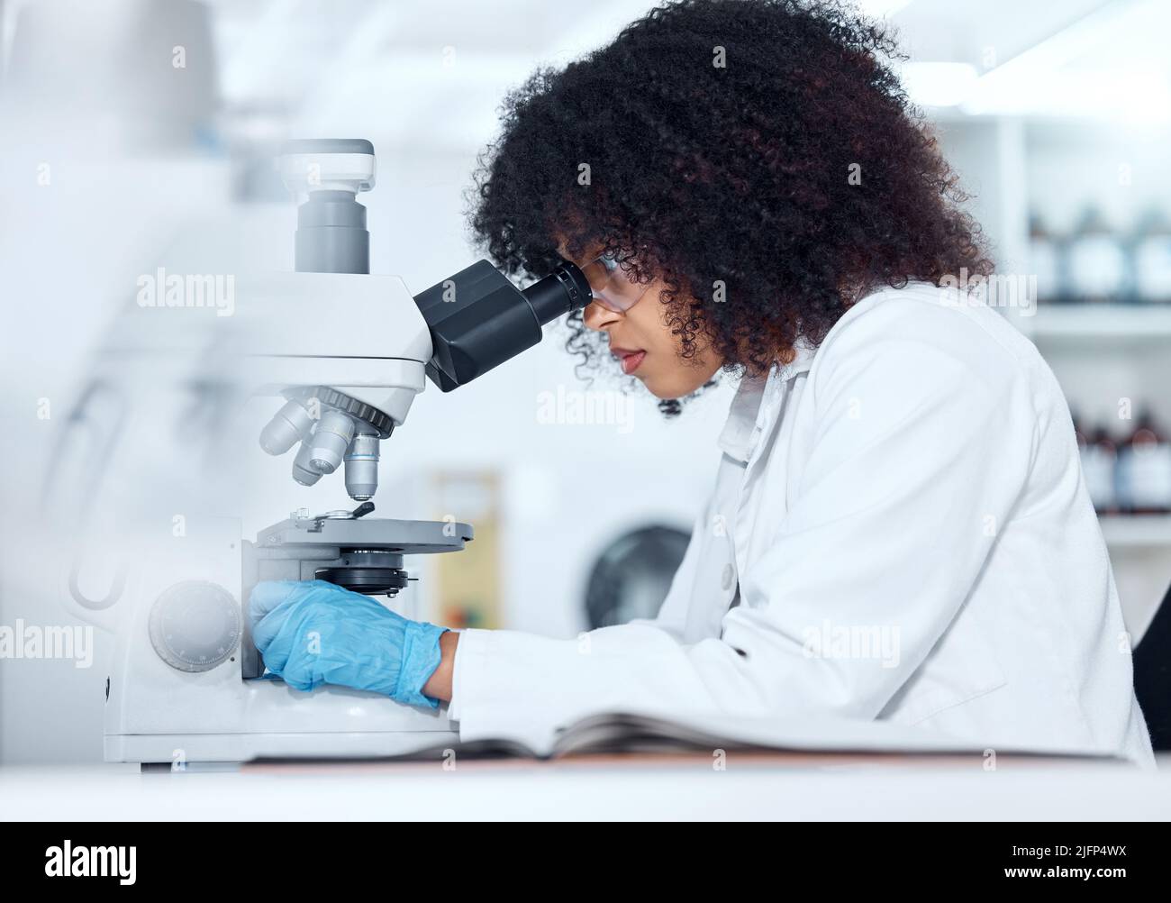 Ein gemischter Rennwissenschaftler mit lockigen Haaren, der Schutzbrillen und Handschuhe trägt und medizinische Testproben in einem Labor auf einem Mikroskop analysiert. Junge Frau tut es Stockfoto