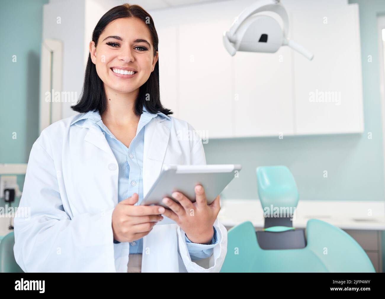 Die junge kaukasische Zahnärztin trägt einen Laborkittel und lächelt, während sie in ihrem Büro ein digitales Tablet benutzt. Zahnhygiene ist wichtig für Ihr Wohlbefinden Stockfoto
