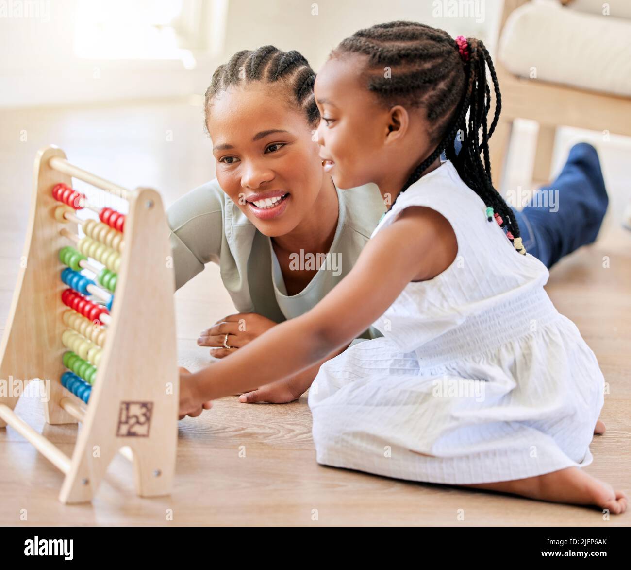 afroamerikanische Mutter, die im Ausfallschritt zu Hause auf dem Holzboden liegt und mit ihrer Tochter lächelt, während sie mit Lernspielzeug spielt. Entzückendes kleines Stockfoto