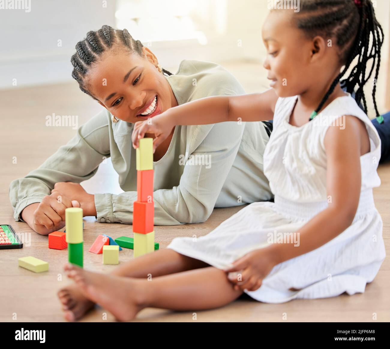 afroamerikanisches Mädchen, das mit Bausteinen spielt, während es mit der Mutter sitzt. Kleines Mädchen baut Turm mit Holzblöcken. Lächelnde Frau, die darauf liegt Stockfoto