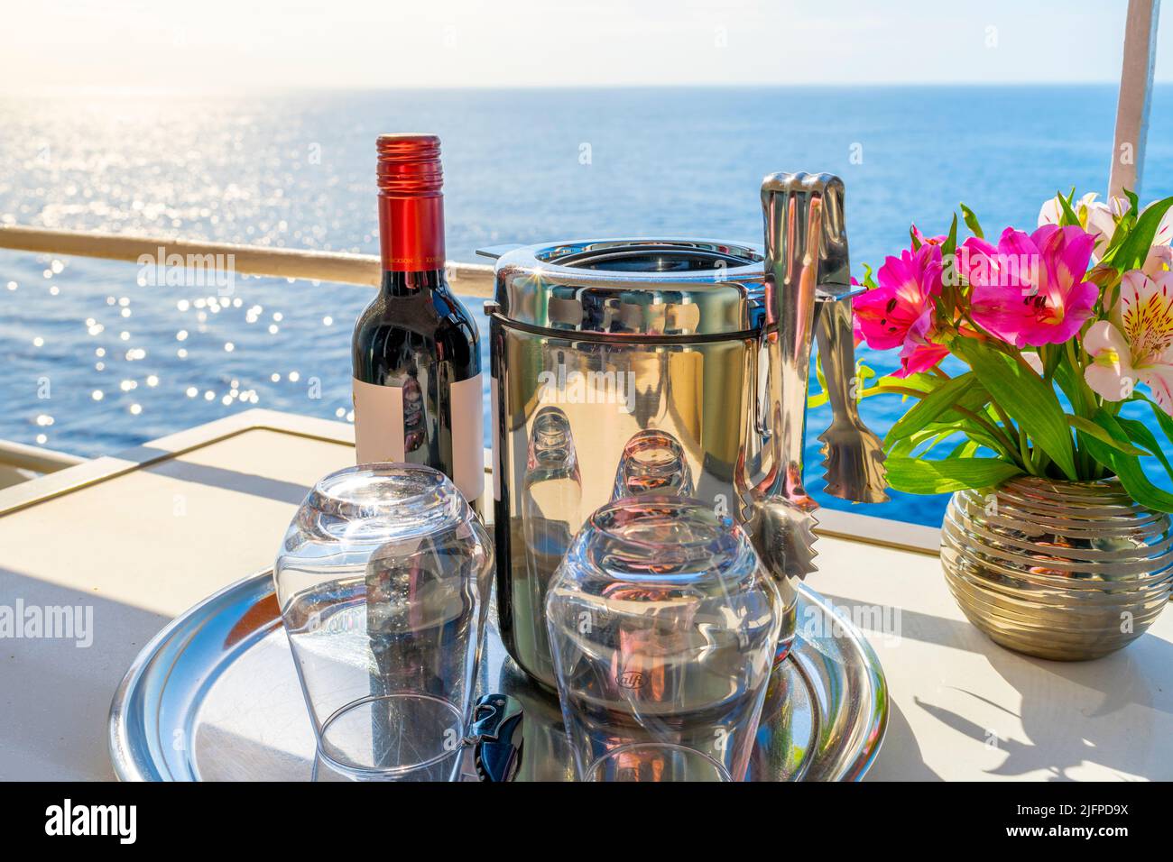 Eine Flasche Rotwein mit Blick auf den Sonnenuntergang, ein Eiskübel und zwei stemless Gläser neben einem Blumenstrauß auf dem Balkon einer Kreuzfahrt-Kabine auf dem Meer. Stockfoto