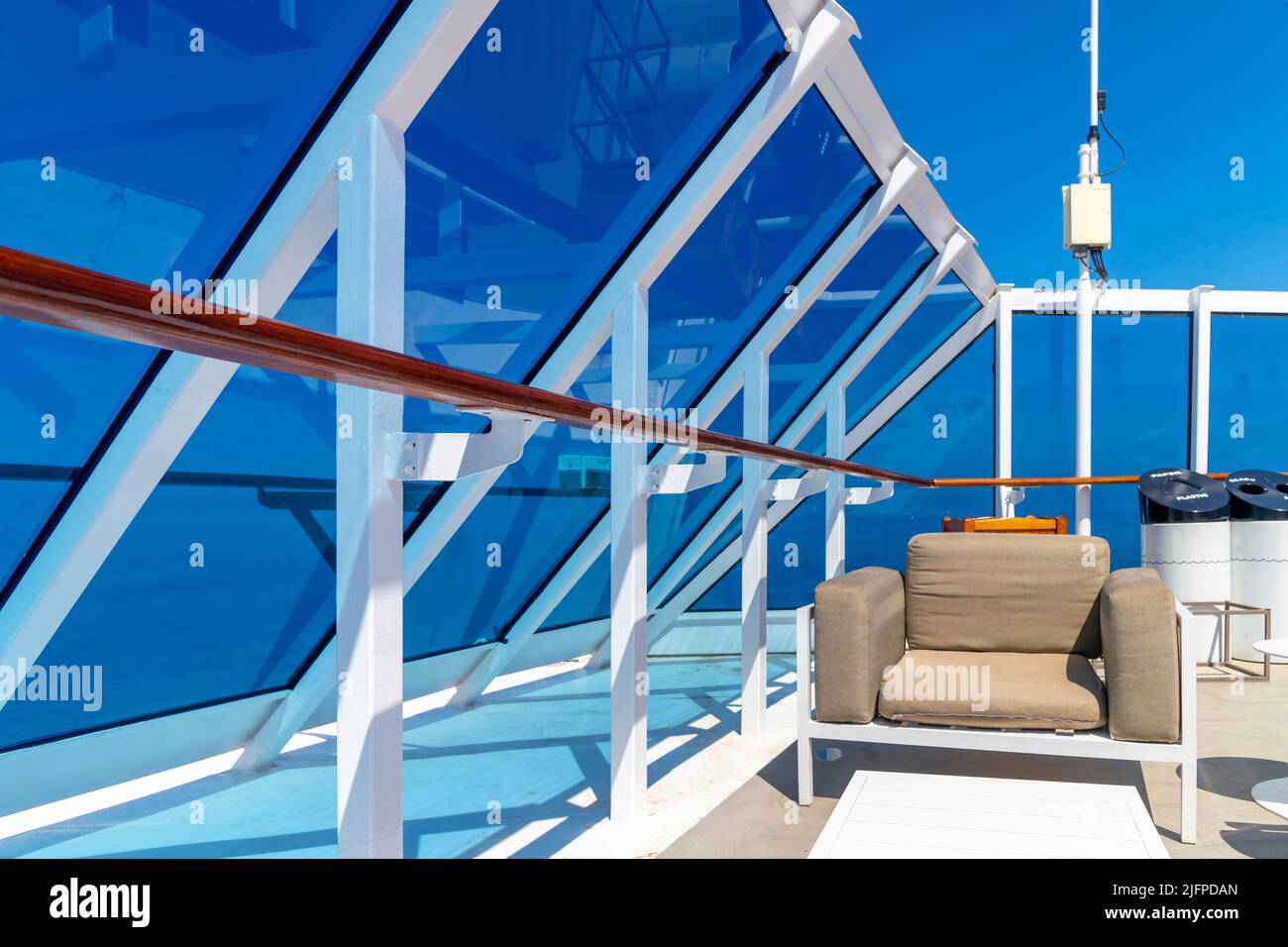Ein Sitzbereich mit Glaspaneelen und weißen Geländern mit tiefblauem Himmel an einem Sommertag auf dem oberen Promenadendeck eines Schiffes auf See. Stockfoto