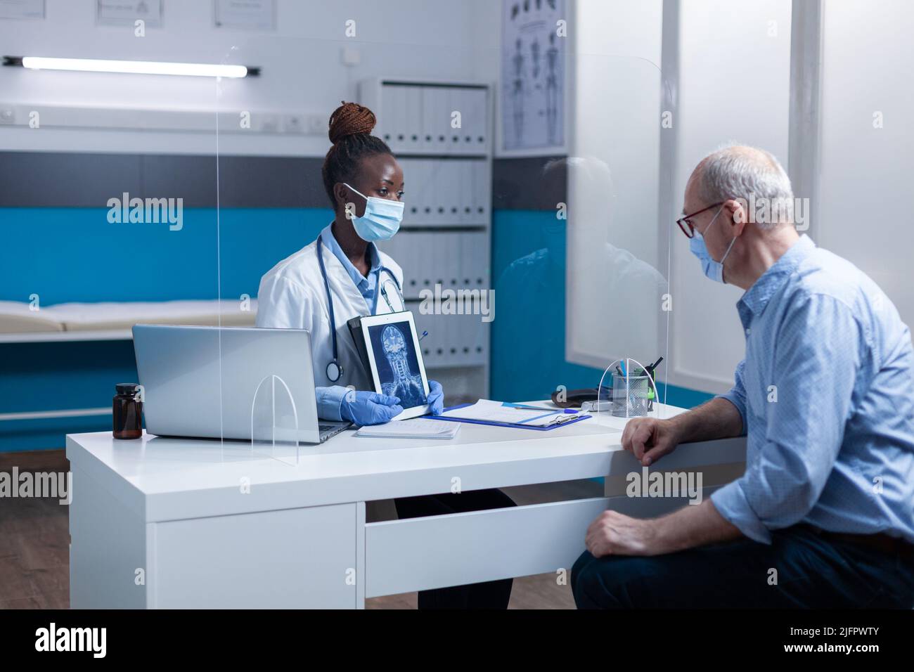 afroamerikanischer Radiologiespezialist erklärt älteren Patienten das Röntgenbild, während sie Gesichtsmasken zum Virenschutz tragen. Ein erfahrener Radiologe zeigt einem älteren Mann ein MRT-Bild. Stockfoto