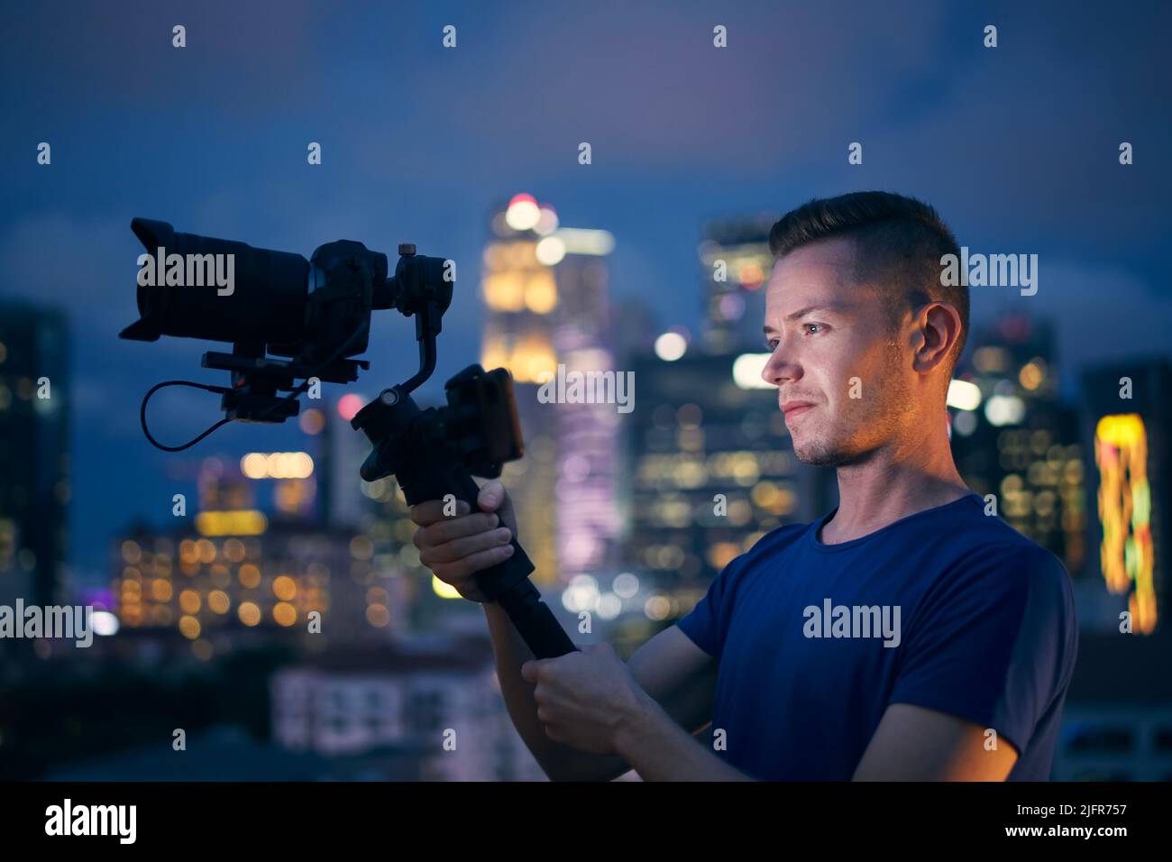 Mann beim Filmen mit Kamera und Gimbal. Porträt eines Videofilmports gegen die beleuchtete Skyline der Stadt bei Nacht. Stockfoto