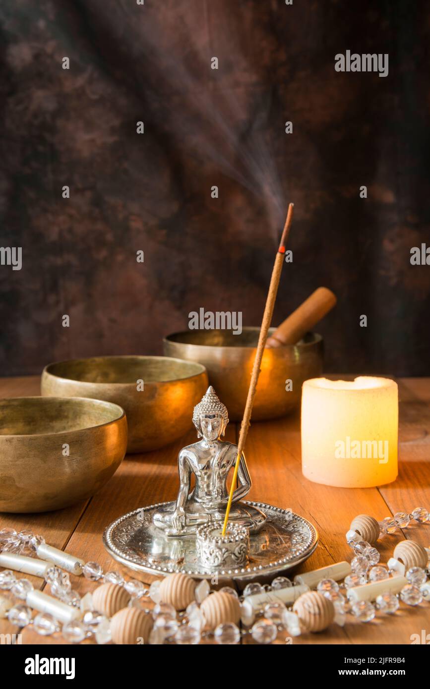 Stillleben mit tibetischen Schalen, einer Perlenkette, einer Kerze, Weihrauch und einer Buddha-Figur auf Holzbrettern und einem dunklen Hintergrund. Kleiner, mit Kerzen angezündete Altar f Stockfoto