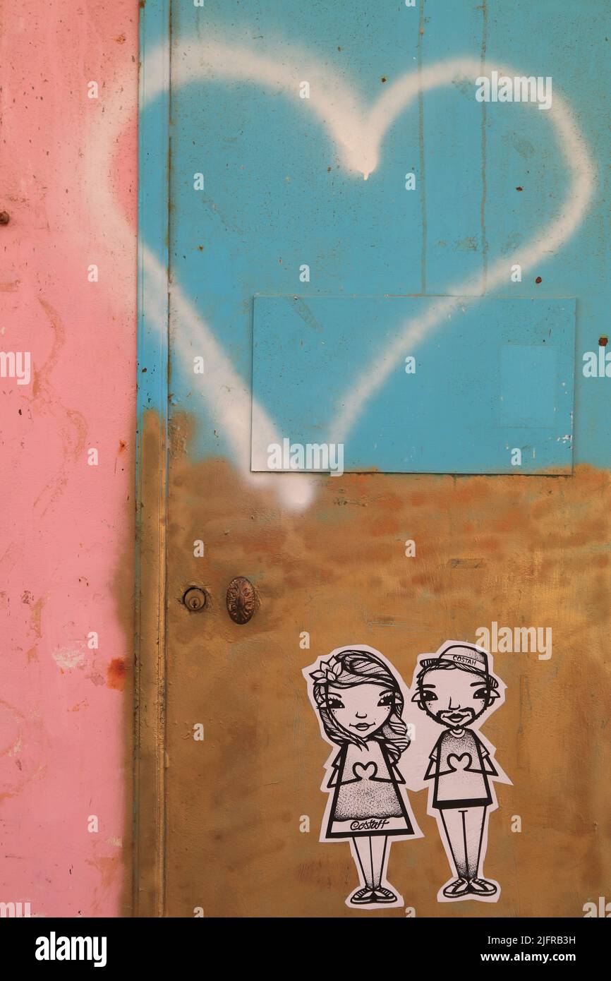 Cartoonisch gezeichnete Charaktere eines Mädchens und Jungen, die ihre Hände in einer Liebe- oder Herzform halten, mit einem großen Herz, das auf einer mehrfarbigen Wand stensiliert ist Stockfoto