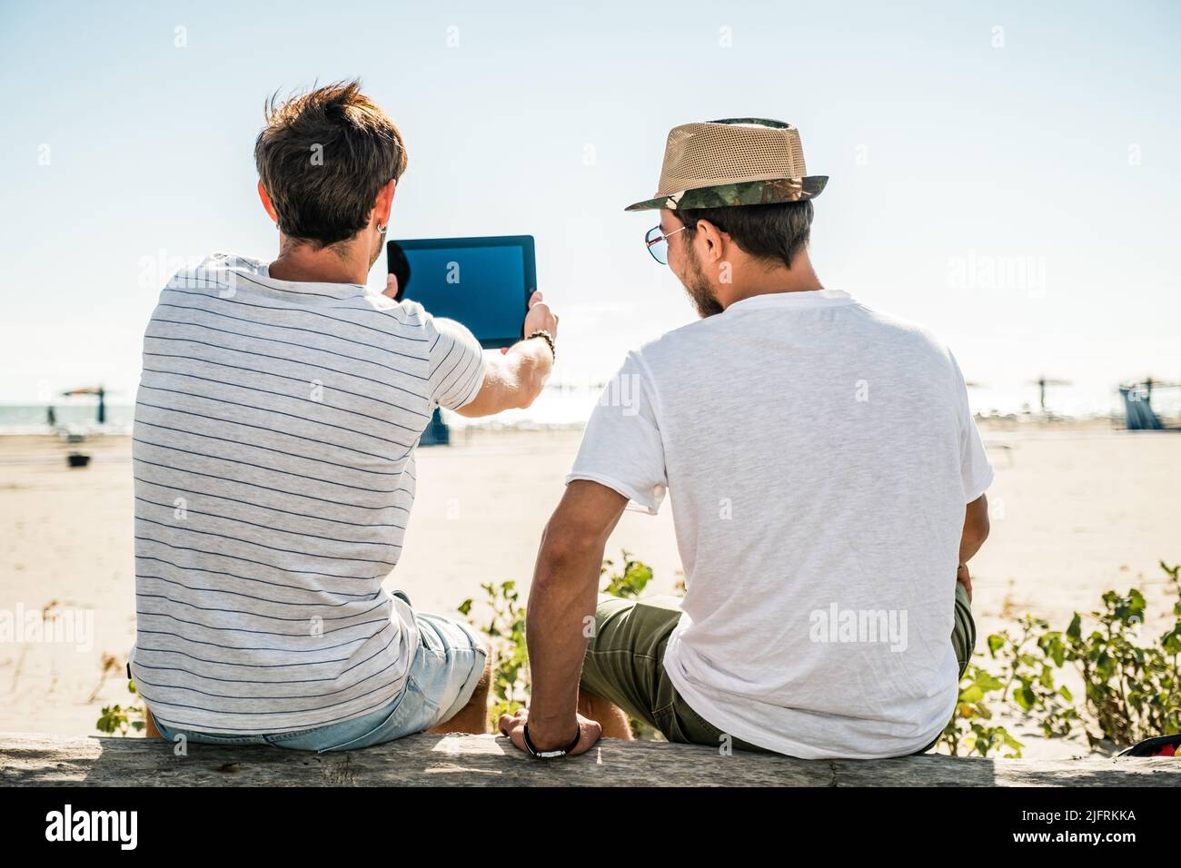 Porträt eines jungen Mannes, der bei Sonnenaufgang mit einem Tablet am Sandstrand sitzt. Reise-, Personen-, Urlaub- und Technologiekonzept Stockfoto