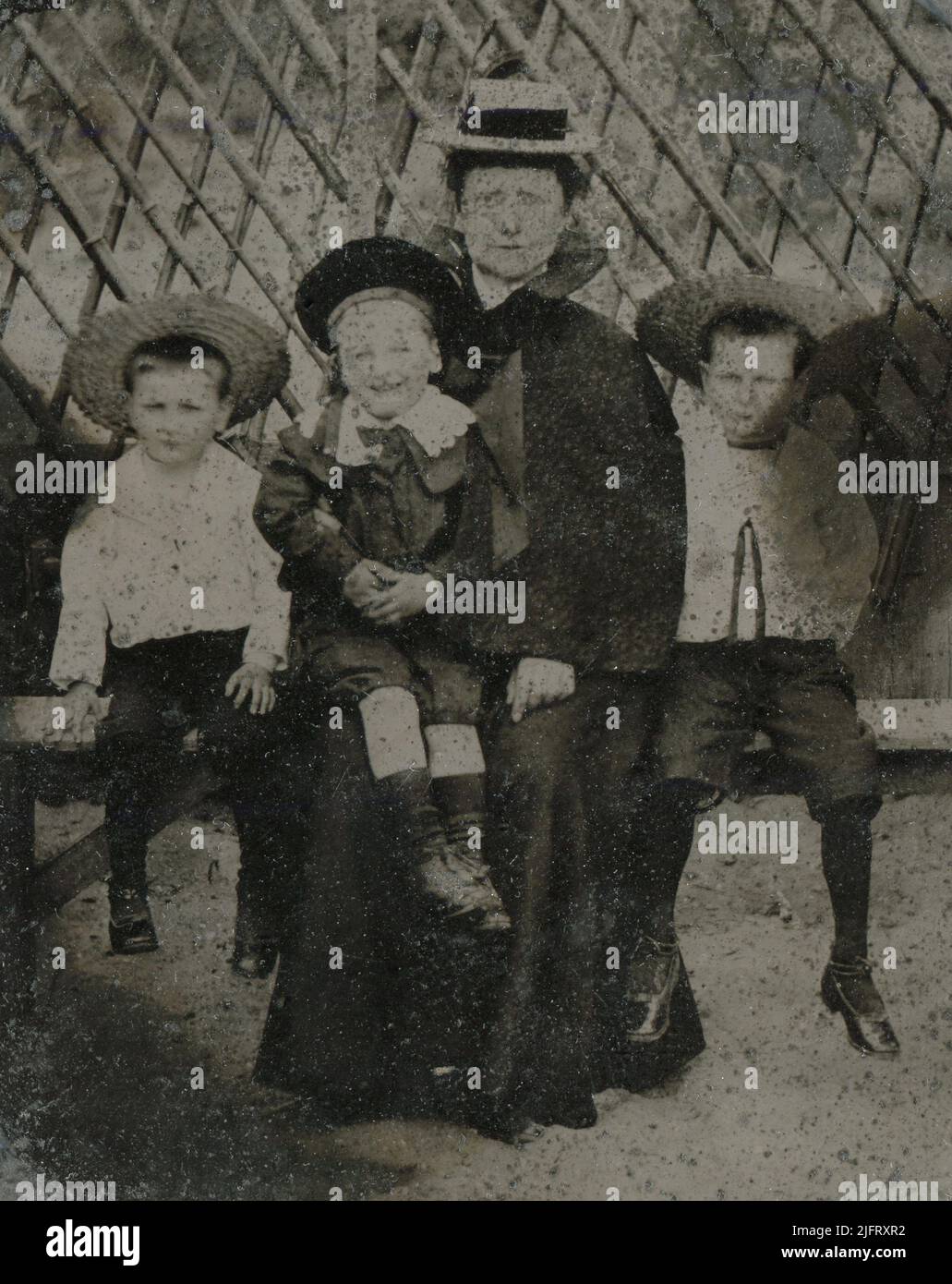 Eine spätviktorianische Porträtaufnahme ‘„Tintype“, auf der eine Mutter mit einem Strohboothut und ihren drei jungen Söhnen auf einer Bank vor einem Gitter sitzt. Zwei der Jungen tragen Matrosenanzüge und Strohhüte, der jüngste einen Seidenanzug mit Knickerbockern, einem Spitzenkragen und einer Tam O' Shanter-Mütze. Stockfoto