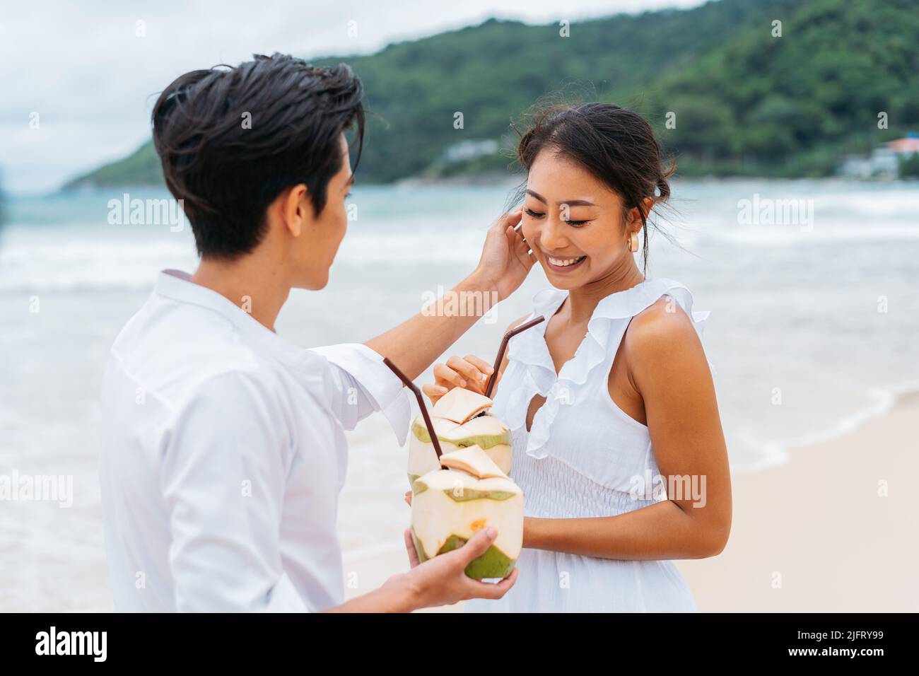 Glücklich lächelnd Asian 30s Paar genießen Reisen auf dem Sandstrand im Sommer. Ein Paar in einem weißen Hemd und Kleid. Liebevolle Liebhaber trinken einen frischen Kokosnusssaft. Stockfoto