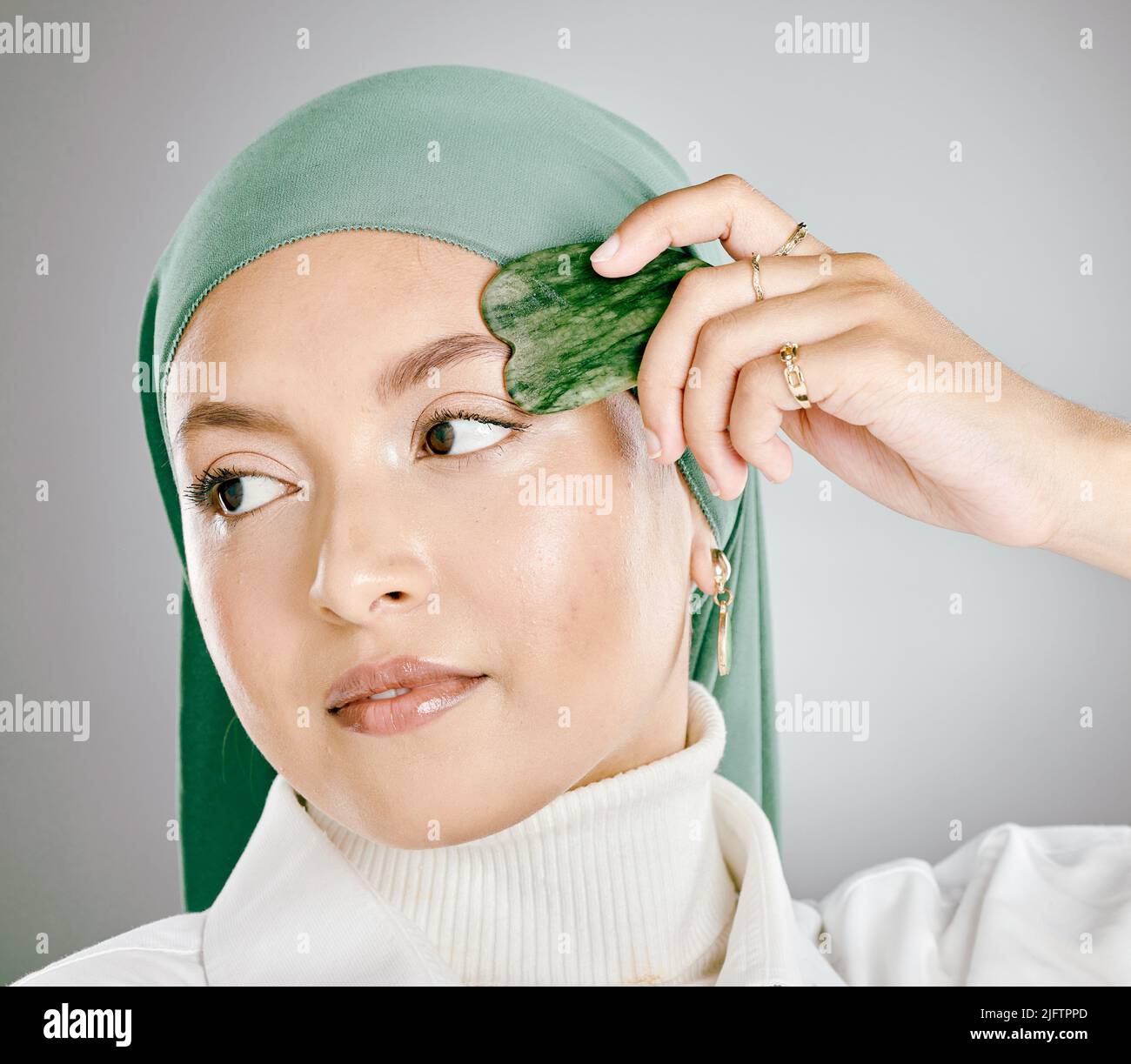 Ein Gua Sha, das auf einem jungen gesicht muslimischer Frauen verwendet wird, isoliert vor grauem Studiohintergrund. Frau mit einem Hijab oder Kopftuch, mit einem Anti-Aging Stockfoto