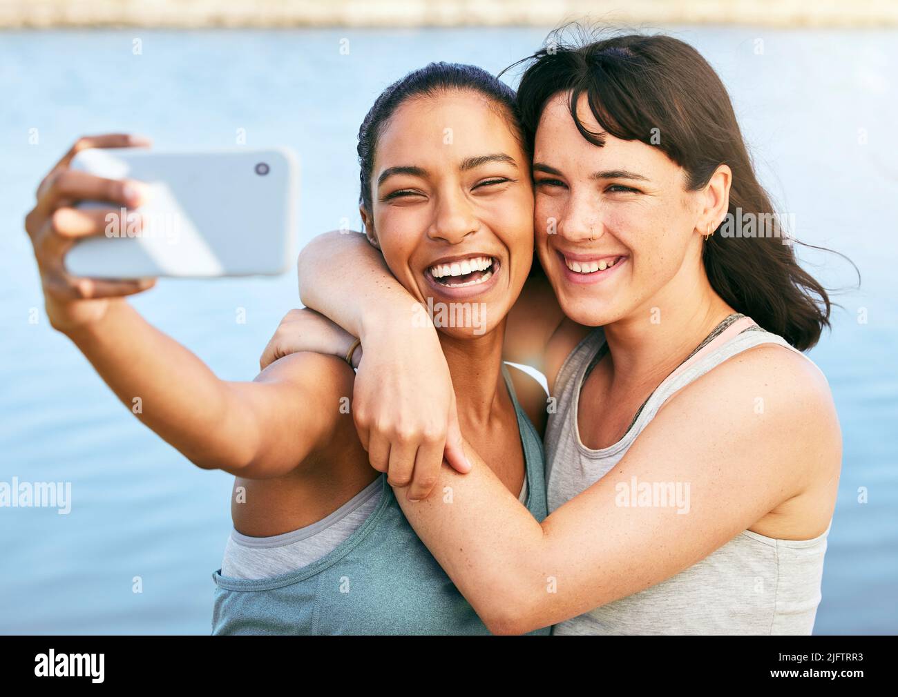 Zwei Freundinnen, die nach einem Training in der Natur Selfies machen und mit einem Smartphone vor einem See stehen. Junge Latino-Hündin, die ihr Telefon benutzt Stockfoto