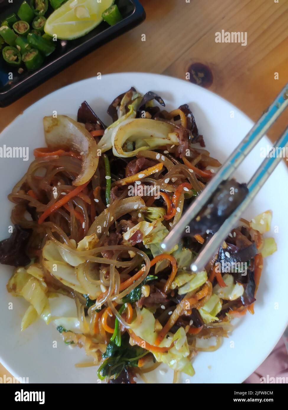 Japchae mit Karotten und Sesam auf dem weißen Teller auf dem Tisch.ist ein herzhaftes und leicht süßes Gericht aus gebratenen Glasnudeln und Gemüse. Stockfoto