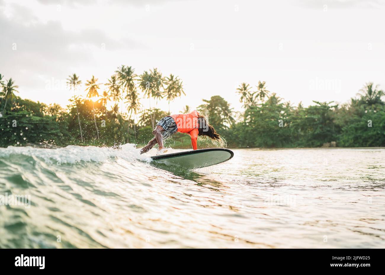 Schwarzer langhaariger Teenager-Junge, der von einem langen Surfbrett reitet und springt. Er fing eine Welle in einer Bucht des indischen Ozeans mit magischem Sonnenuntergang Hintergrund. Extreme Wa Stockfoto