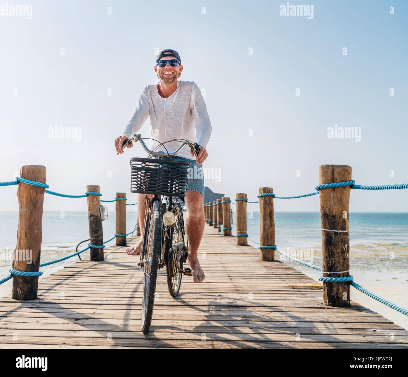 Porträt eines glücklichen lächelnden Mannes, gekleidet in leichte Sommerkleidung und Sonnenbrille, der auf dem hölzernen Seebrücke mit dem Fahrrad fährt. Sorgloser Urlaub im tropischen Stockfoto