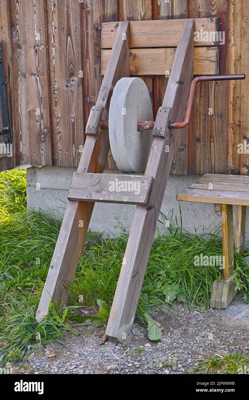 Ein antiker Schleifstein, der gegen einen Holzschuppen mit überwuchertem Gras steht. Altes graues Schärfsteinwerkzeug oder Ausrüstung, die draußen auf einem Bauernhof verwendet wird Stockfoto