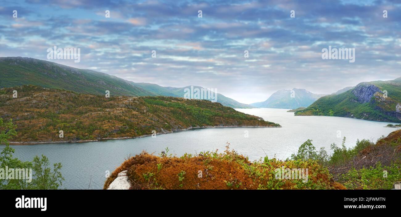 Flusslandschaft zwischen Hügeln und Bergen. Grünes Laub am Flussufer mit blauem Himmel in Norwegen. Ruhiges Wasser in der Nähe einer lebhaften Wildnis Stockfoto