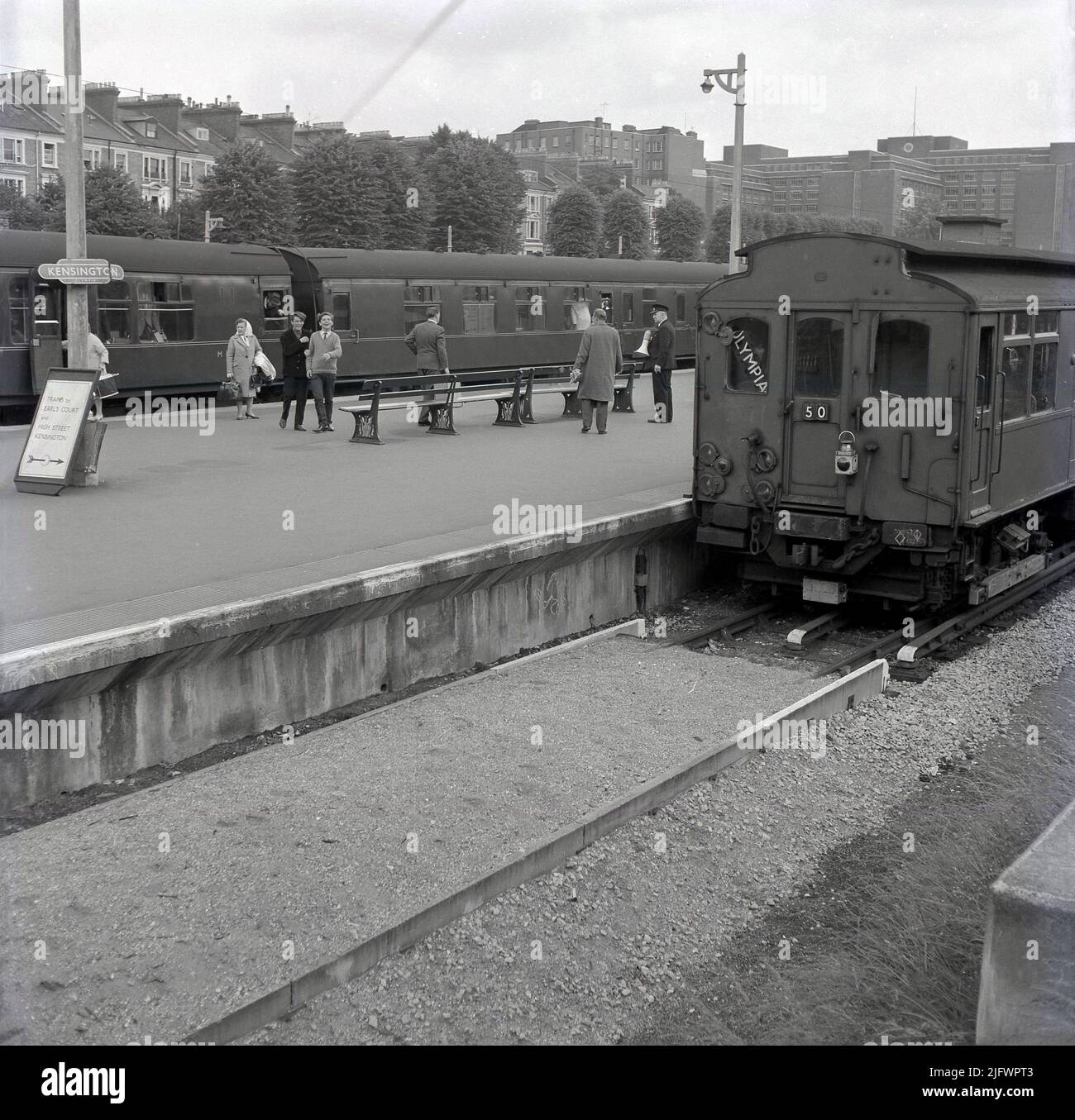 1960s, historische Ansicht von Kensington Olympia Bahnhof und U-Bahnstation, Kensington, London, England. VEREINIGTES KÖNIGREICH. Ein Hauptzug wartet auf dem Bahnsteig, während ein Schild sagt, dass Züge nach Earls Court und High Street Kensington auf der rechten Seite sind, wo eine oberirdische U-Bahn wartet, mit der Welt Olympia im Fenster. Stockfoto