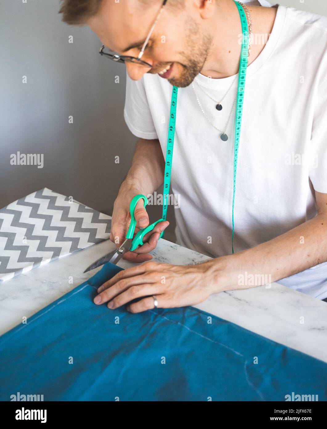 Ein lächelnder Schneider schneidet Stoff in einem Heimstudio. Ein autodidaktischer Seemann in weißem T-Shirt und Brille arbeitet mit Stoff, Muster und Kreide. Stoffschnitt Stockfoto