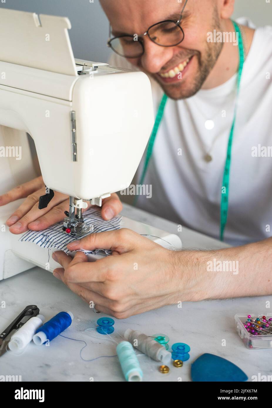 Ein lächelnder, blonder Mann in einer Brille näht in der heimischen Werkstatt. Schneiderin mit einem Maßband am Hals in einem weißen T-Shirt. Das Konzept des kleinen Unternehmens Stockfoto