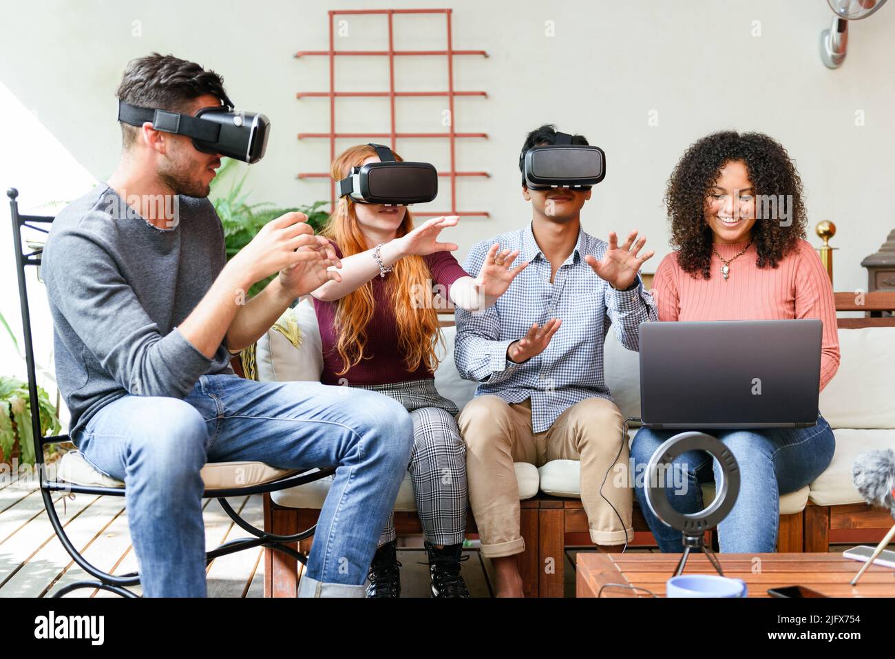 Multirassische Männer und Frauen in legerer Kleidung mit VR-Helmen interagieren mit dem Cyberspace, während sie in der Nähe ihrer Freundin sitzen und auf dem Laptop in der Sonne spielen Stockfoto
