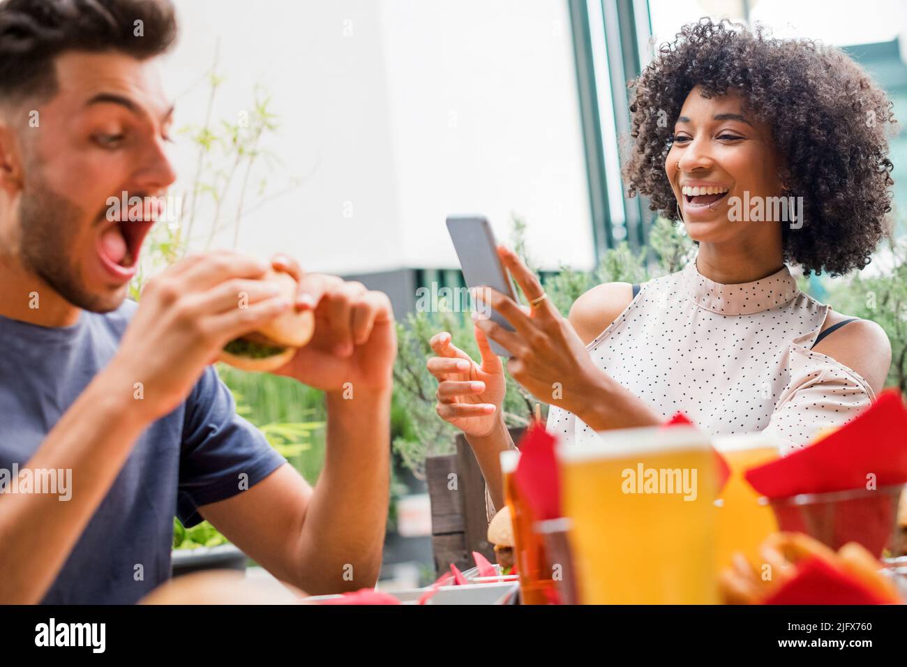 Das junge Paar genießt es, an einem Restauranttisch Fast Food zu essen und lacht, als sie Medien auf ihrem Handy lesen Stockfoto
