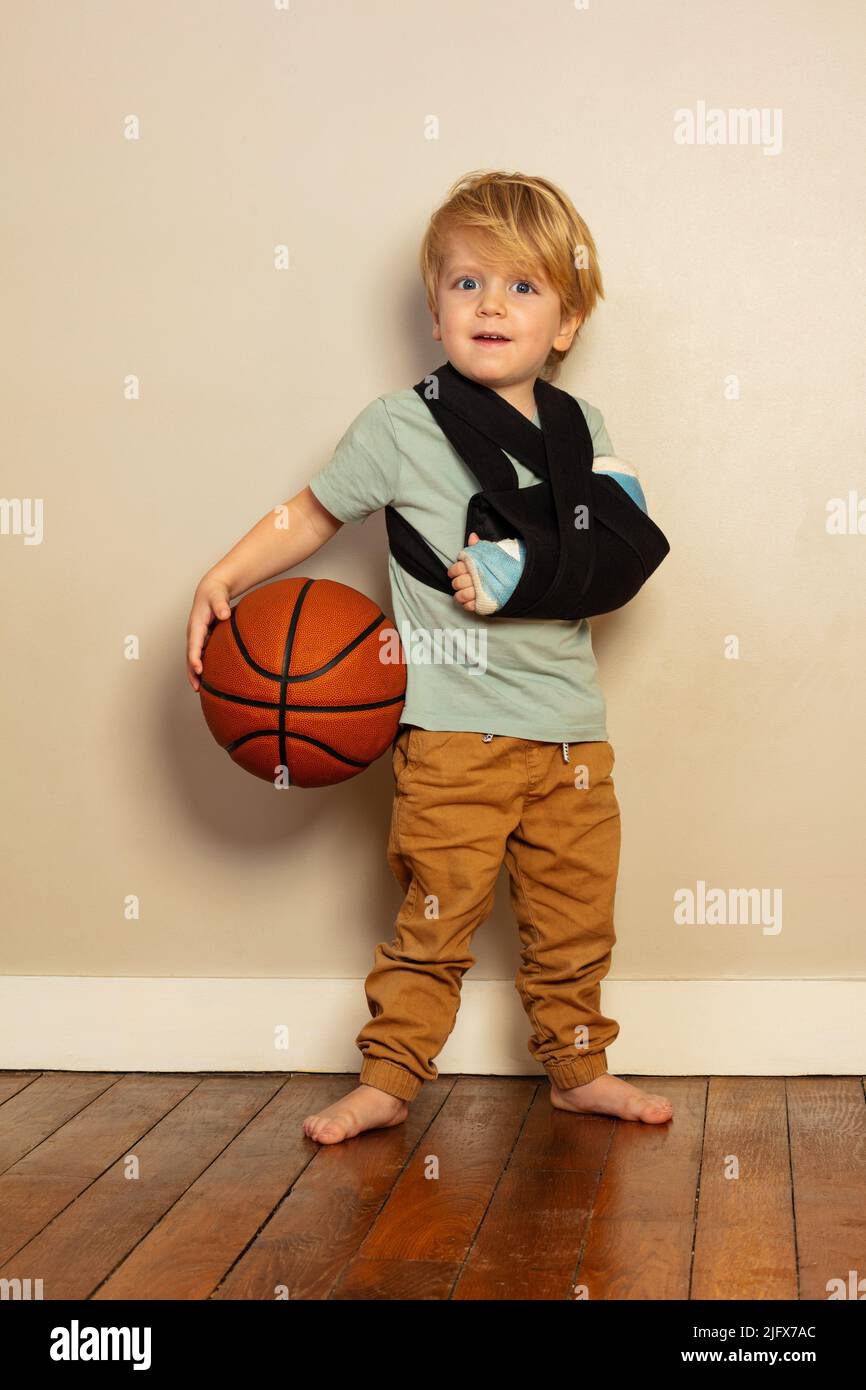 Kleiner blonder Junge mit gebrochener Hand, der den Basketballball hält Stockfoto