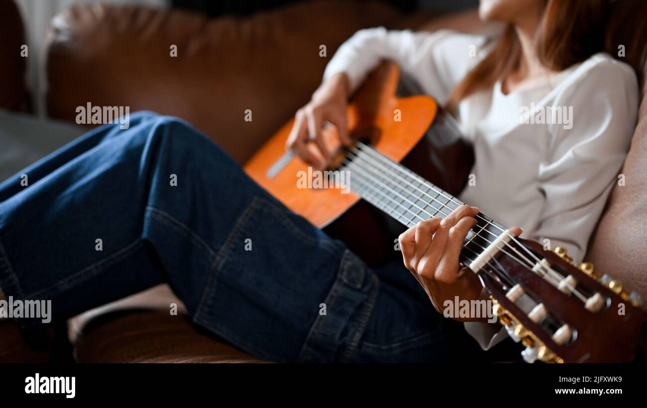 Talentiertes und ruhiges junges asiatisches Mädchen, das auf dem Sofa liegt und im Wohnzimmer eine klassische Gitarre spielt. Kreatives Hobby-Konzept. Zugeschnittenes Bild Stockfoto