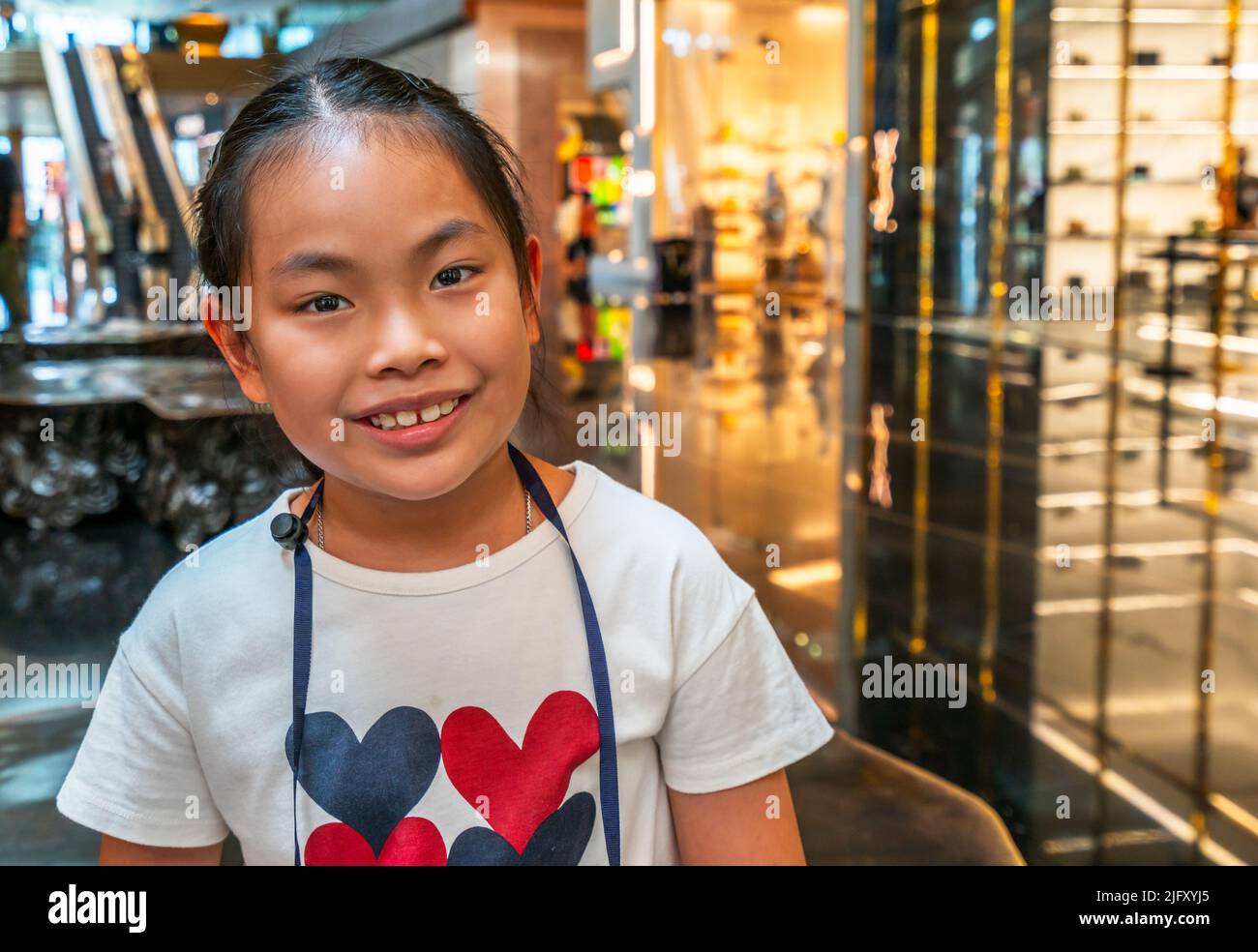 Porträt von niedlichen asiatischen Mädchen in einem Einkaufszentrum, lächelndes Gesicht, Nahaufnahme Porträt des Kindes, Augen Blick auf die Kamera, verschwommener Hintergrund des Einkaufszentrums Stockfoto