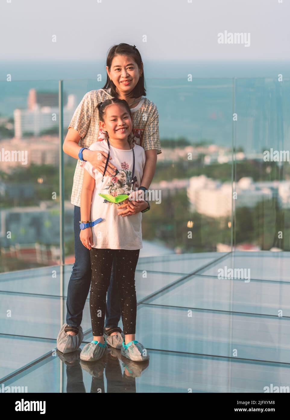 Glücklich und aufgeregt stehen die asiatische Mutter mittleren Alters und die kleine Tochter auf einem Glasboden auf einem hohen Gebäude, voller Körper, lächelndes Gesicht, verschwommener Rücken Stockfoto