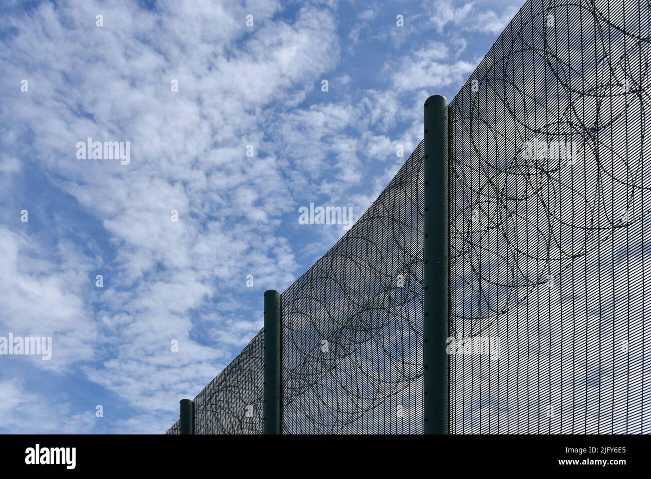 Sicherheitszaun aus Metall mit Rasierdraht auf der Oberseite gegen einen blauen Himmel mit weißen Wolken. Stockfoto
