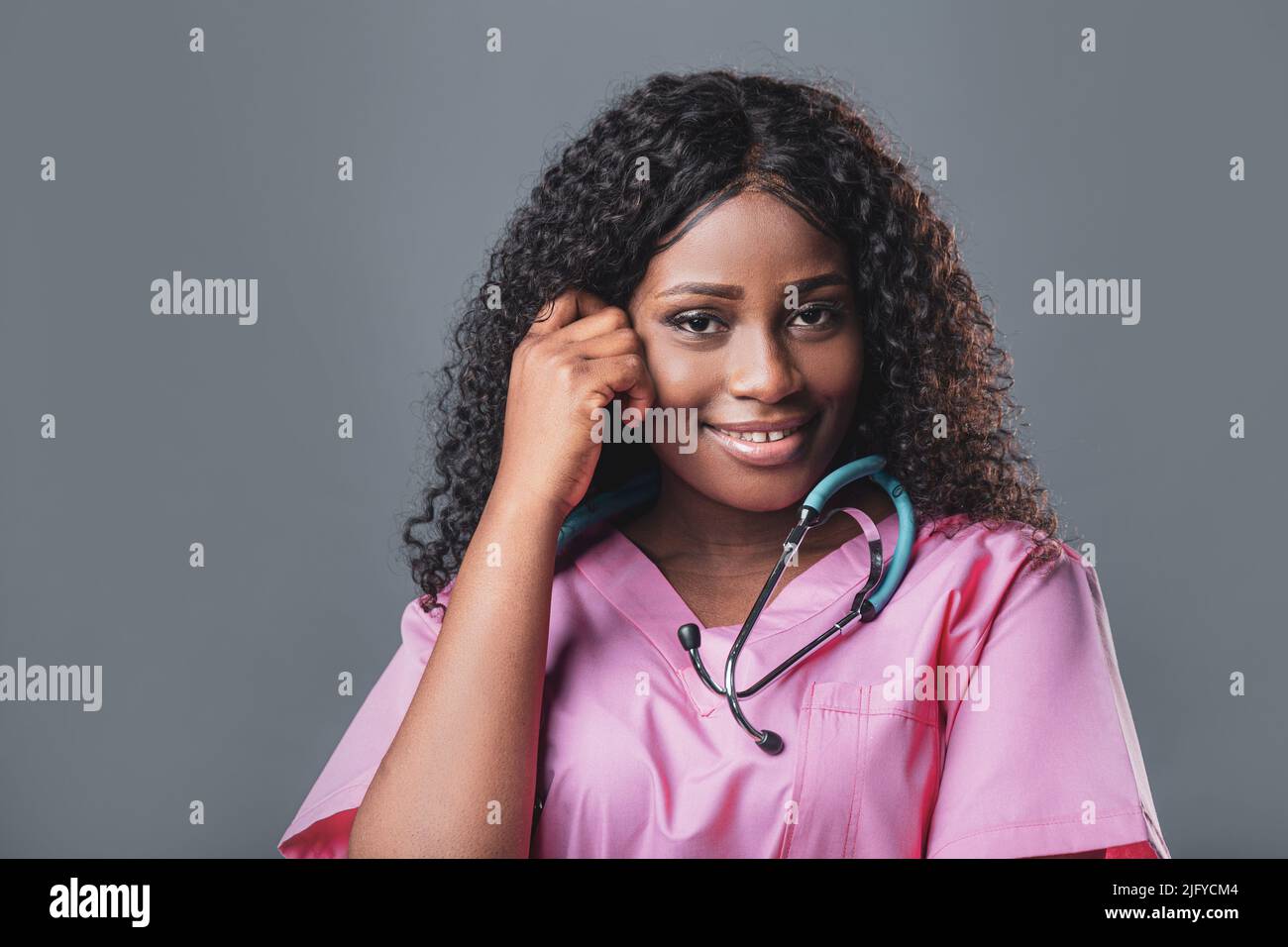 Junge Ärztin schwarz afrikanische Frau lächelt mit Stethoskop in rosa Uniform auf grauem Hintergrund. Stockfoto