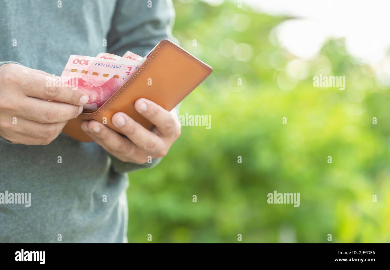 Asiatischer Mann zählt und überprüft chinesische Banknoten in seiner Brieftasche. Außenaufnahmen mit grün verwacklungsunscharfer Hintergrundfarbe Stockfoto