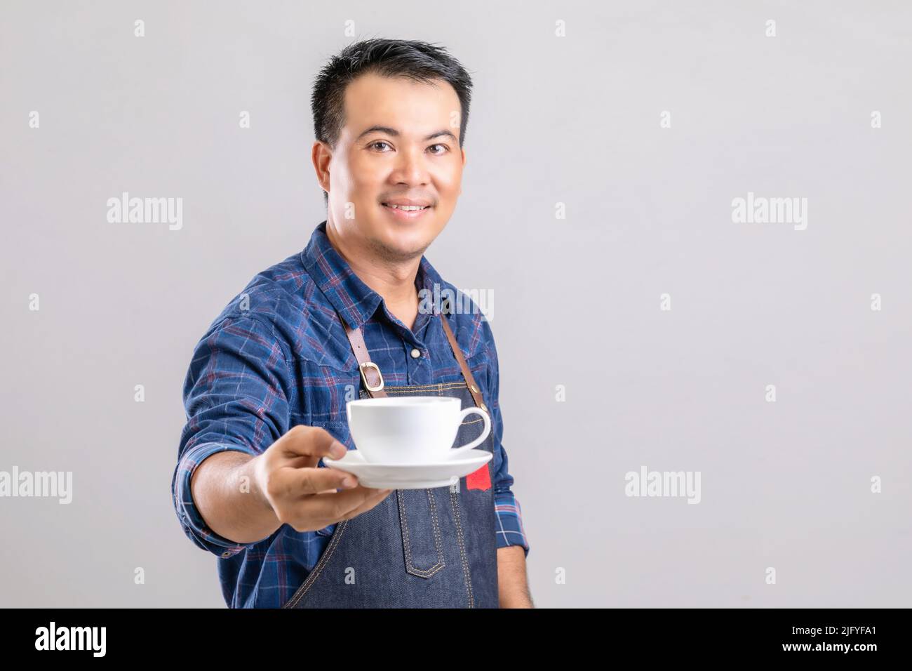 Portrait Asian Mann in Barista Uniform hält weißen Kaffeetasse isoliert auf grauem Hintergrund Stockfoto