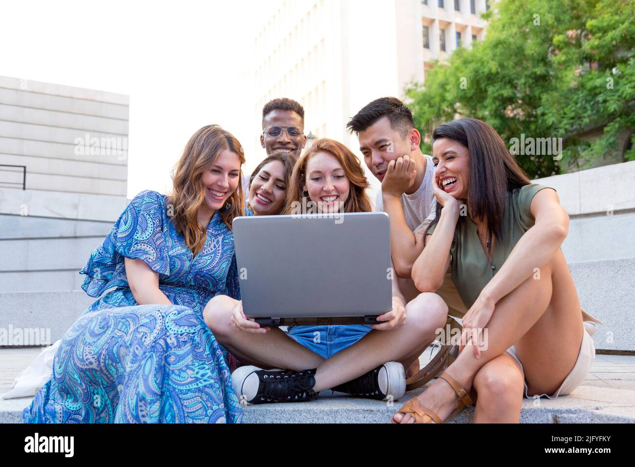 Eine Gruppe multiethnischer Freunde, die auf einem Stadtpark sitzen, während sie zusammen einen Laptop benutzen Stockfoto