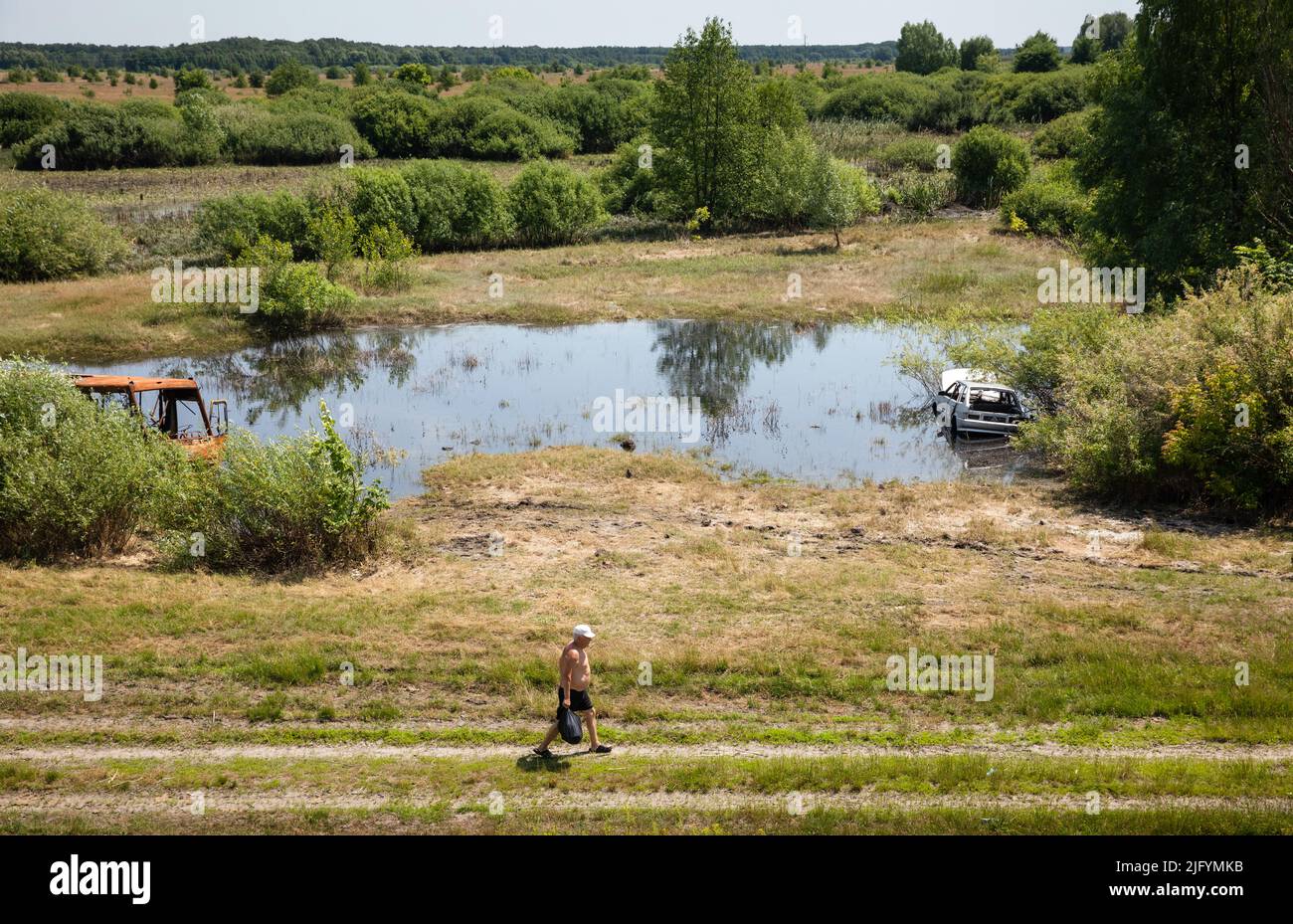 TSCHERNIHIW REG, UKRAINE - 19. Juni 2022: Krieg in der Ukraine. Ein einsamer Mann geht mit einem ausgebrannten Bus und einem zerstörten Auto auf einem Weg in der Nähe eines Sees um sein Geschäft Stockfoto