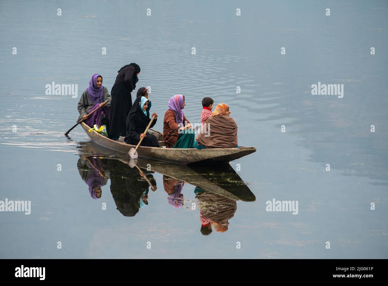 Srinagar, 10, April, 2016; Reflexion des kleinen Bootes mit Kashmiri Familie in der traditionellen Tracht, die Dal See kreuzt, Srinagar, Kashmir, Indien Stockfoto