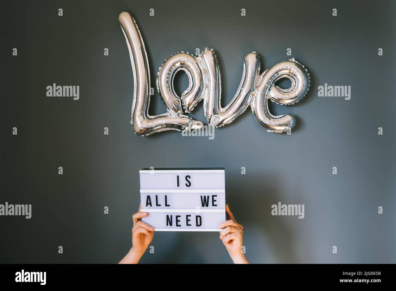 Männliche Hände halten Leuchtkasten mit Text Love ist alles, was wir brauchen unter silbernen Folienballon auf grauen Wand Hintergrund. Tonung des Filmeffekts. Valentinstag Konz Stockfoto