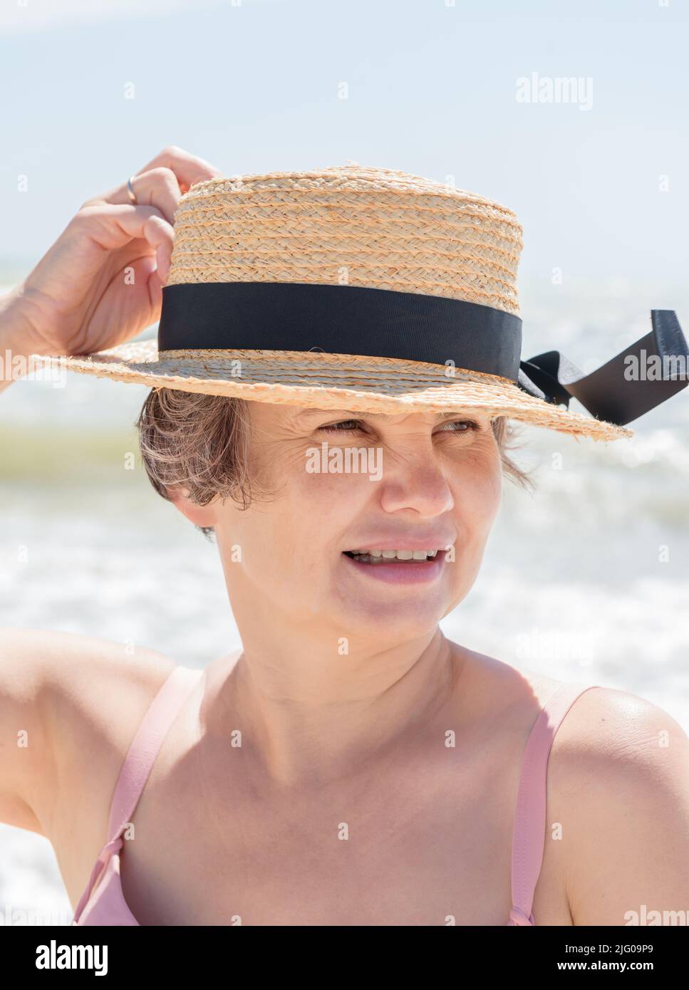 Verheiratete grauhaarige Frau in Bootsanzug und Badeanzug, die ihren Hut vor der Meeresbrise auf verschwommenem Meeresgrund mit Wellen stützte.Konzept der Weiblichkeit. Stockfoto