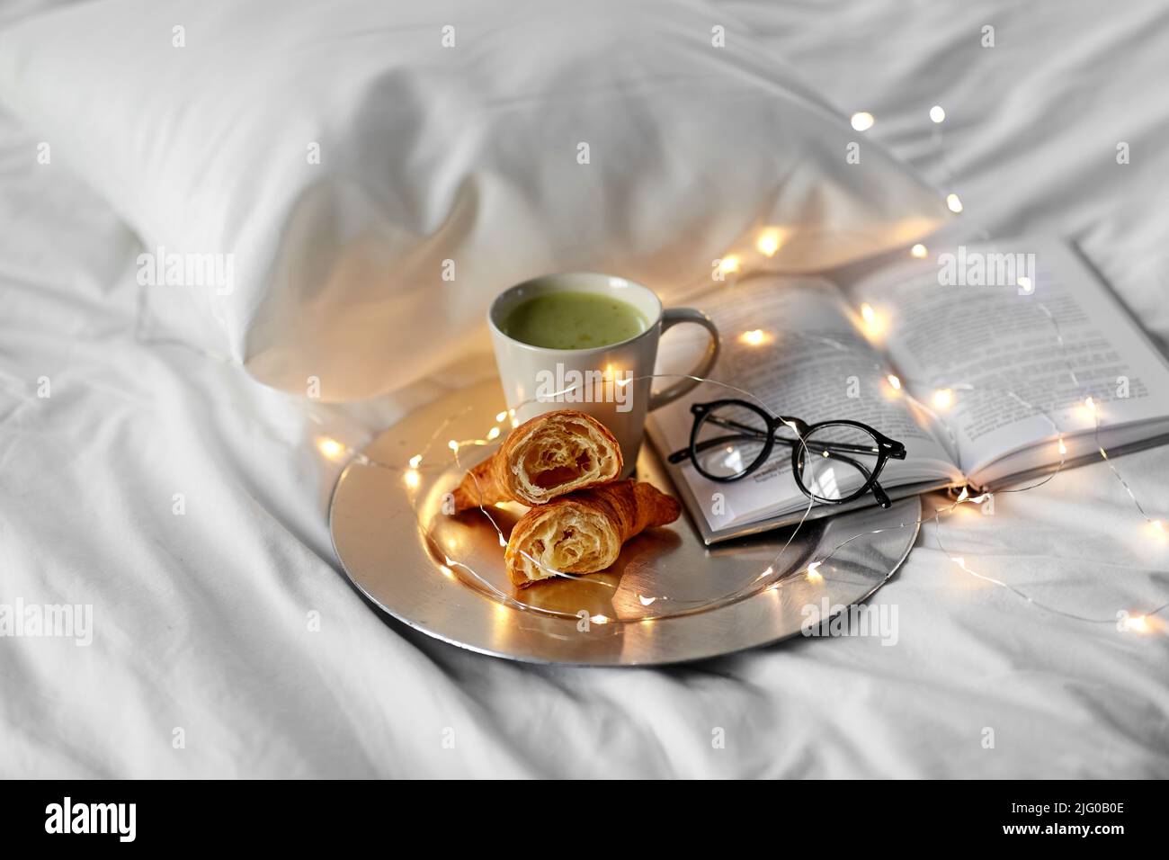 Croissants, Matcha-Tee, Buch und Gläser im Bett Stockfoto