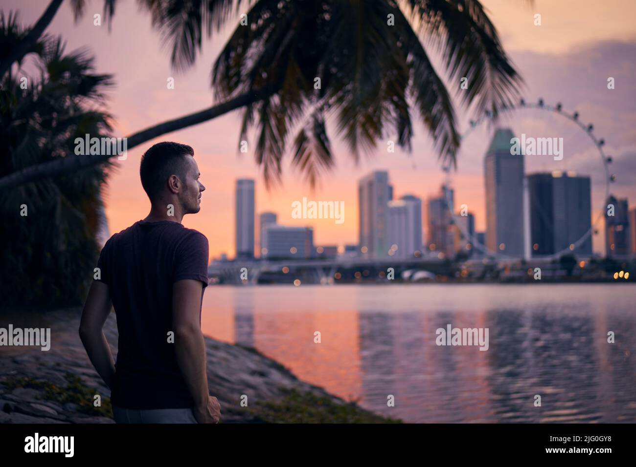 Ein hübscher Mann, der unter einer Palme am Ufer der Bucht steht und die Stadt anschaut. Skyline der Stadt bei Sonnenuntergang, Singapur. Stockfoto