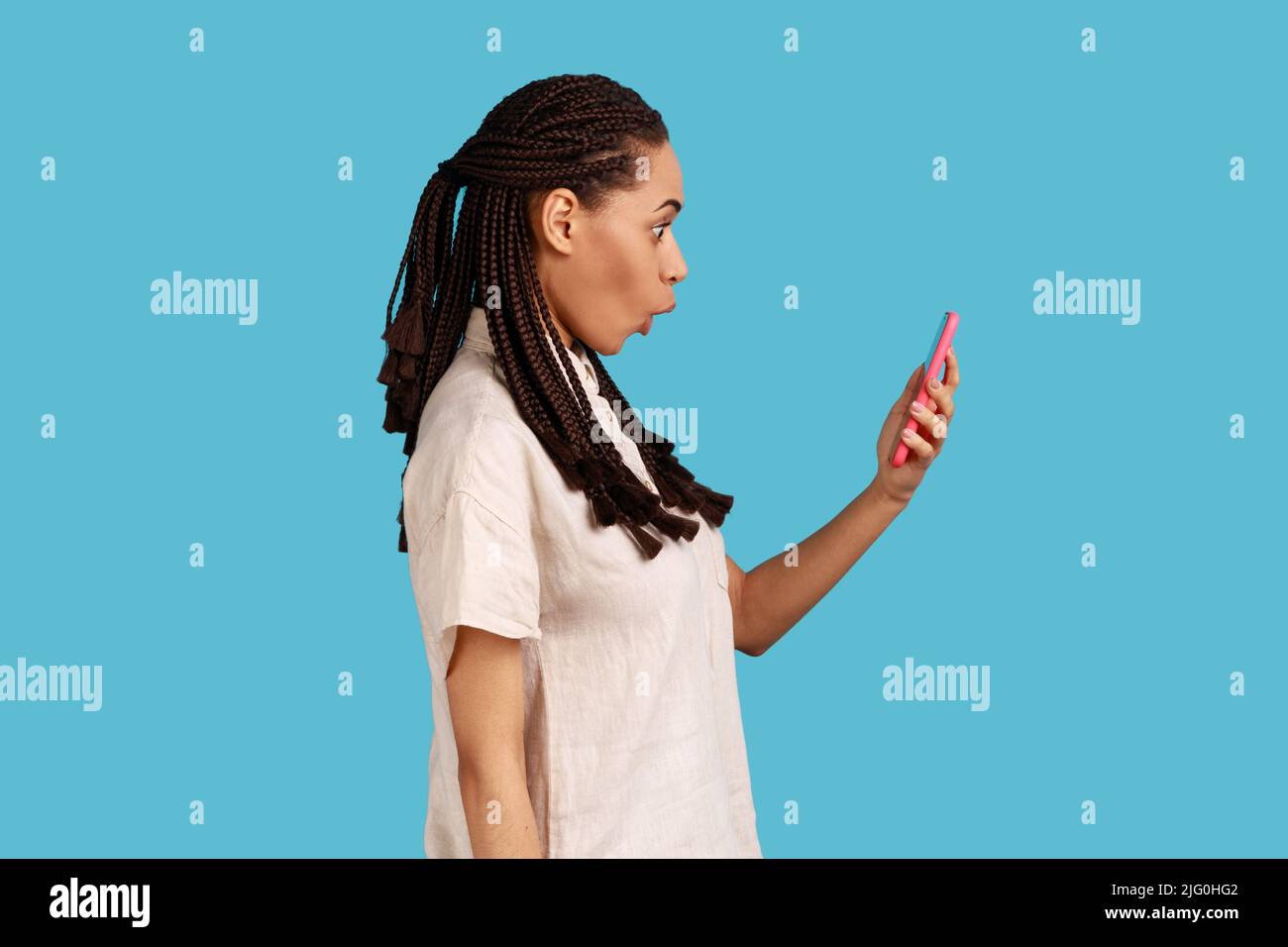 Seitenansicht der überraschten Frau mit schwarzen Dreadlocks starrt auf den Smartphone-Bildschirm, liest erstaunliche Nachrichten mit erstauntem Gesicht, von etwas fassungslos. Innenaufnahme des Studios isoliert auf blauem Hintergrund. Stockfoto
