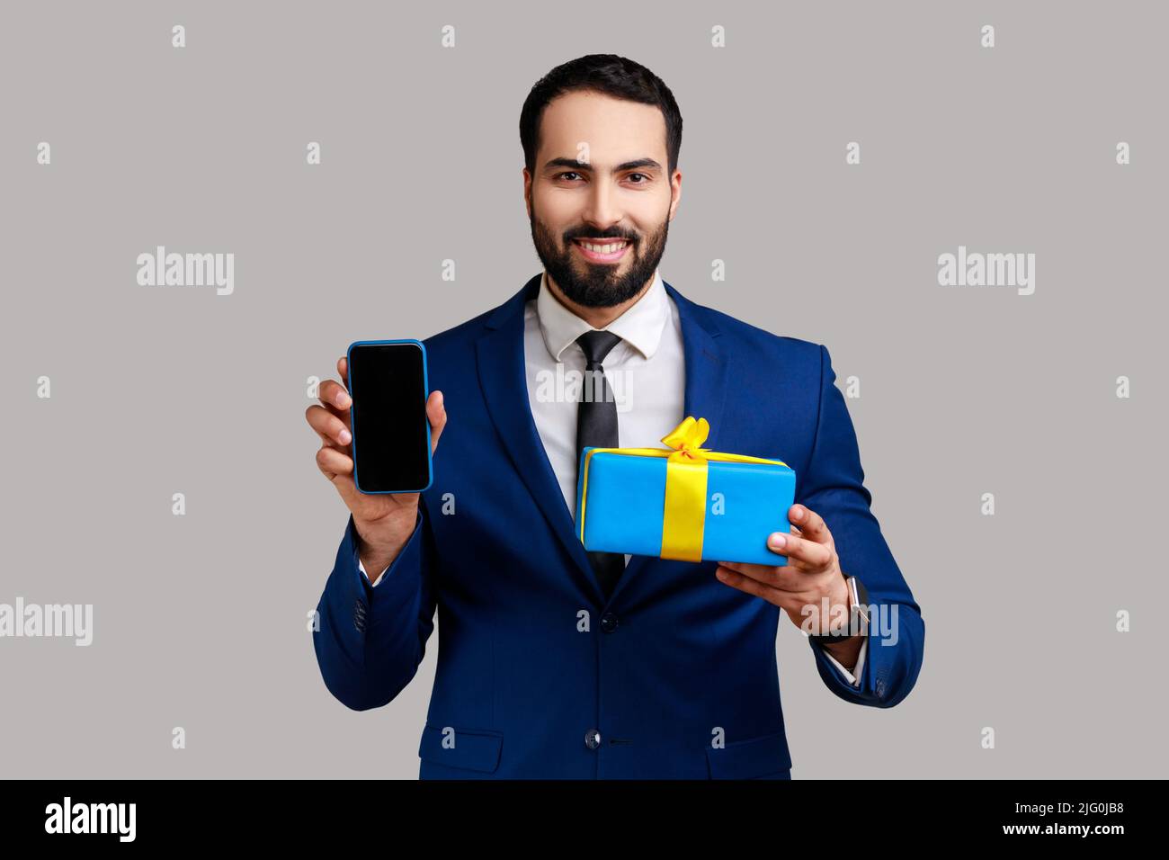 Bärtiger Geschäftsmann hält Geschenkbox und Handy mit leerem Display für Online-Shopping-Werbung und trägt einen offiziellen Style-Anzug. Innenaufnahme des Studios isoliert auf grauem Hintergrund. Stockfoto