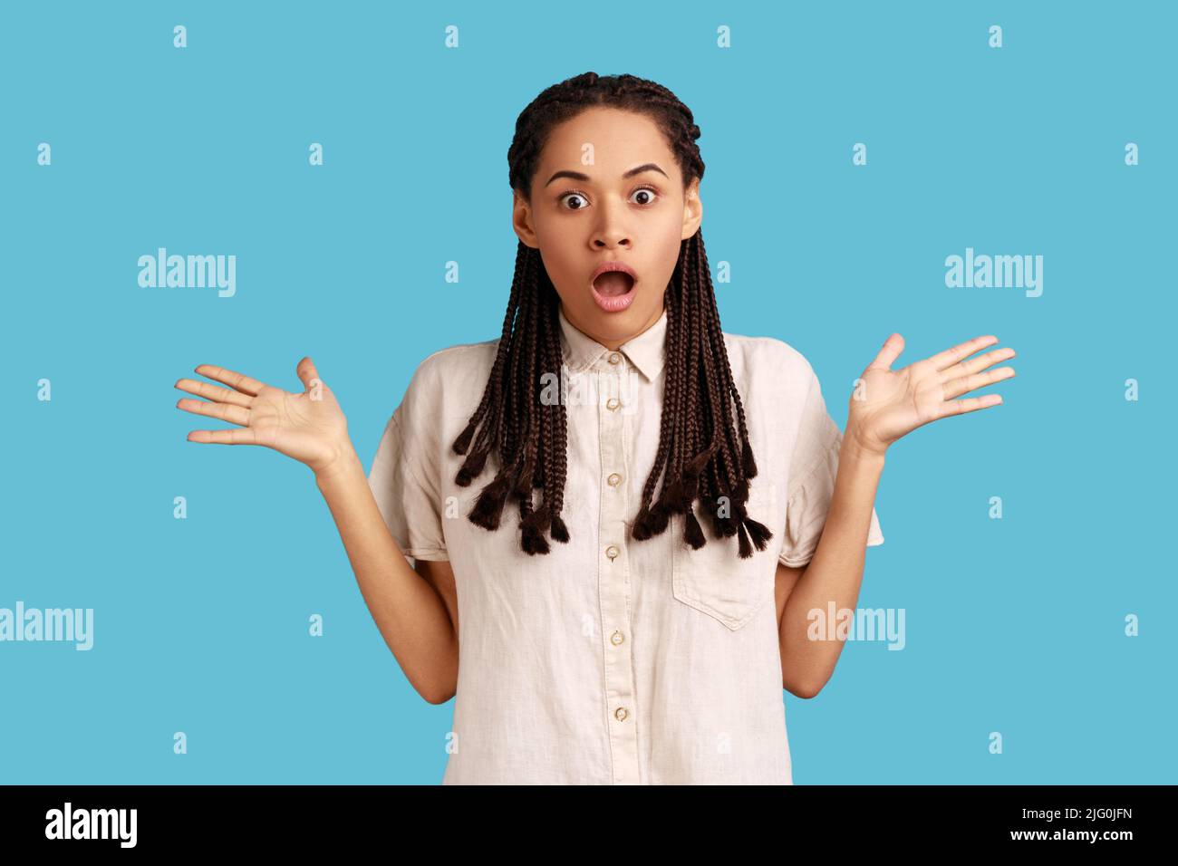 Empört schockiert Frau mit schwarzen Dreadlocks hat wütend Ausdruck überrascht, hob die Arme, Blick auf die Kamera mit großen Augen, trägt weißes Hemd. Innenaufnahme des Studios isoliert auf blauem Hintergrund. Stockfoto